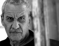 Schwarz-weiß Porträt eines älteren Mannes mit lockigem Haar und ernstem Ausdruck. Er steht neben einer unscharfen Holzstruktur.