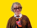 Eine Person mit Brille und grauen Haaren lächelt, trägt eine bunte Krawatte und einen braunen Pullover vor einem gelben Hintergrund.