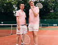 Zwei Männer in rosa Poloshirts und weißen Shorts stehen auf einem Tennisplatz mit Schlägern. Im Hintergrund ist ein Netz zu sehen.