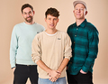Drei Männer stehen vor beigem Hintergrund. Sie tragen lässige Kleidung und lächeln in die Kamera.