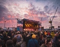Menschenmenge bei einem Konzert im Freien, vor einer beleuchteten Bühne bei Sonnenuntergang. Kräne sind im Hintergrund zu sehen.