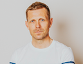 Ein Mann mit kurzem Haar, trägt ein weißes T-Shirt. Er blickt ernst in die Kamera vor einem neutralen Hintergrund.