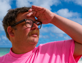 Mann mit Brille und Schnurrbart im rosa Shirt hält die Hand über die Augen, um in die Ferne zu schauen, blauer Himmel im Hintergrund.