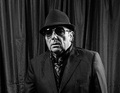 Mann in Anzug, Hut und Sonnenbrille steht vor einem dunklen Vorhang, schaut ernst in die Kamera. Schwarz-Weiß-Foto.