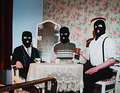 Drei Personen in schwarzen Skimasken sitzen an einem Tisch mit Spitzen-Tischdecke in einem Raum mit floraler Tapete.