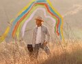 Eine Person mit Cowboyhut steht in einem Feld, im Hintergrund bunte, gemalte Regenbögen.