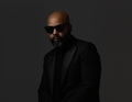 Mann mit Glatze und Vollbart trägt einen dunklen Anzug und Sonnenbrille, posiert vor dunklem Hintergrund.