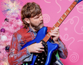 Mann spielt blaue E-Gitarre vor rosa Hintergrund mit glitzerndem Weihnachtsbaum. Er trägt eine bunte Jacke und schaut konzentriert.