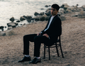Ein Mann im Anzug sitzt auf einem Stuhl am steinigen Ufer. Im Hintergrund ist das Wasser sichtbar.