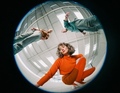 Drei Personen in moderner Kleidung, fisheye Perspektive von unten. Eine Person hockt, zwei stehen, helle Architektur im Hintergrund.