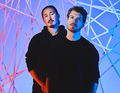Zwei Männer in schwarzen Hoodies stehen nebeneinander vor einem geometrischen, blau-rot beleuchteten Hintergrund.