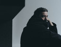 Person in dunkler Kleidung lehnt nachdenklich an einer Wand, schaut zur Seite. Atmosphäre ist dunkel und minimalistisches Design.