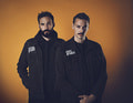 Zwei Männer in dunkler Kleidung stehen vor einem orangefarbenen Hintergrund. Beide haben kurze Haare und ernste Gesichtsausdrücke.