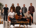 Acht Männer posieren mit Musikinstrumenten, darunter eine Gitarre und ein Akkordeon, in einem Studio vor einem Sofa.