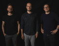 Drei Männer stehen vor dunklem Hintergrund, zwei lächeln. Sie tragen dunkle Kleidung und blicken entspannt in die Kamera.