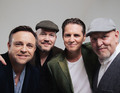 Vier Männer posieren lächelnd nebeneinander. Zwei tragen Mützen, einer trägt ein weißes Hemd, der andere einen grünen Blazer.