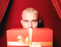 Eine Person mit blonden Haaren schaut über eine große rote Geschenkbox mit gelber Schleife, vor einem roten Hintergrund mit Vorhängen.