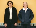 Zwei Männer stehen vor einer gelben Wand. Einer trägt einen schwarzen Blazer, der andere eine dunkle Jacke. Beide lächeln leicht.
