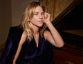 Eine Frau mit blonden Haaren lehnt sich nachdenklich an einen Flügel, in eleganter dunkler Kleidung.