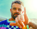Ein Mann mit Bart zeigt eine Handgeste. Er trägt ein buntes, gehäkeltes Oberteil und steht vor einem hellen, verschwommenen Hintergrund.