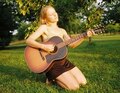 Eine Frau kniet im Gras und spielt Gitarre. Sie schaut verträumt nach oben. Im Hintergrund sind Bäume zu sehen.