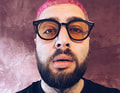 Person mit pinken Haaren, Vollbart und runden Brillengläsern vor rosafarbenem Hintergrund, trägt ein schwarzes Oberteil.