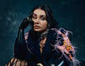 Frau mit dunklem Haar trägt schwarzen Handschuh und Jacke mit Federn. Sie lehnt leicht nach vorne vor dunklem Hintergrund.