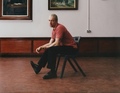 Ein Mann in rotem T-Shirt sitzt seitlich auf einem Stuhl in einem Kunstgalerie-Raum, Bilder an der Wand im Hintergrund.