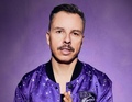Ein Mann mit kurzem Haar und Schnurrbart trägt eine lila Jacke vor einem lila Hintergrund.