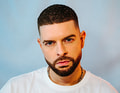 Ein Mann mit kurzem, dunklem Haar und Bart trägt ein weißes T-Shirt. Der Hintergrund ist sanft blau.