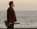 Ein Mann steht seitlich am Meer mit einer Gitarre in der Hand, blickt in die Ferne.