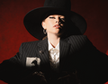 Person in einem schwarzen Nadelstreifenanzug und großem Hut, steht vor rotem Hintergrund. Licht- und Schatteneffekt betont Gesicht.