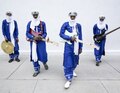 Vier Männer in blauen traditionellen Gewändern und weißen Tüchern spielen elektrische Gitarren und ein Schlaginstrument vor weißer Wand.