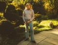 Eine Person steht auf einem gepflasterten Weg in einem sonnigen Garten mit geschnittenen Büschen und trägt ein gestreiftes Hemd sowie Jeans.