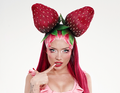 Person mit pinkem Haar, trägt ein Kopfschmuck aus zwei großen Erdbeeren und schaut direkt in die Kamera.