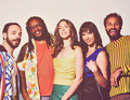 Eine Gruppe von fünf Personen steht lächelnd nebeneinander vor einem neutralen Hintergrund, farbenfroh gekleidet.
