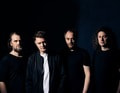 Vier Männer in schwarzer Kleidung stehen nebeneinander vor einem dunklen Hintergrund. Sie blicken ernst in die Kamera.
