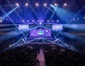 Eine Bühne in einer großen Halle, beleuchtet mit bunten Scheinwerfern und mehreren Bildschirmen, umgeben von einem großen Publikum.