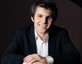 Ein lächelnder Mann in einem dunklen Anzug und weißem Hemd sitzt vor einem dunklen Hintergrund, die Hände vor sich gefaltet.