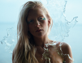 Eine Frau mit langem blondem Haar steht vor verschwommenem Hintergrund, umgeben von transparenten, wasserähnlichen Strukturen.