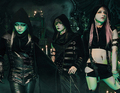 Drei Personen in dunkler, gothischer Kleidung stehen in einem schummrigen Raum mit Kerzen und grünlichem Licht.