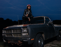 Eine Person sitzt auf der Motorhaube eines alten, dunkelgrauen Trucks in einer nächtlichen Umgebung.
