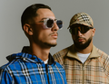 Zwei Männer in karierter Kleidung; einer trägt eine Sonnenbrille, der andere eine Baseballkappe. Sie stehen vor grauem Hintergrund.