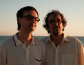 Zwei Männer mit Sonnenbrillen stehen vor einem Meer im Sonnenuntergang, tragen helle Hemden und schauen nachdenklich in die Ferne.