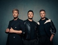 Drei Männer, die lässig posieren, tragen dunkle Kleidung vor einem neutralen Hintergrund.