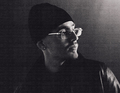 Schwarz-weiß Bild einer Person mit Brille und Mütze, die zur Seite schaut. Die Szene ist dunkel beleuchtet.