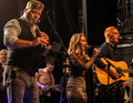 Eine Band tritt auf: Ein Mann spielt Flöte, eine Frau singt, ein anderer Mann spielt Gitarre. Bühne mit Beleuchtung.