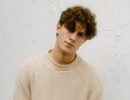Junger Mann mit lockigem Haar trägt beigefarbenen Pullover, steht vor einer weißen, leicht abblätternden Wand und blickt ernst.