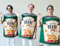 Drei Personen in Baked Beans-Kostümen winken und lächeln; sie tragen weiße Hosen und stehen vor einem neutralen Hintergrund.