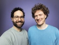 Zwei lächelnde Männer vor einem lila Hintergrund. Der eine trägt Brille und graues Sweatshirt, der andere ein blaues Sweatshirt.